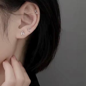 Könnte beinhalten: Nahaufnahme eines Ohrs mit drei silbernen Initialenohrringen. Die Ohrringe buchstabieren die Buchstaben "A", "C" und "S".