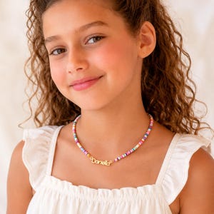 May include: A colorful beaded necklace with the name "Daniella" in gold lettering. The necklace features a variety of pastel-colored beads. The necklace is worn by a person wearing a white top.