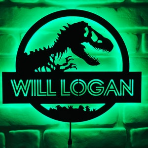 May include: A black dinosaur silhouette with the text "WILL LOGAN" illuminated in green. The design is set against a green-lit background, mounted on a brick wall. The image features a circular design with a dinosaur and the name.
