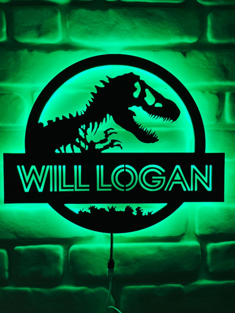 May include: A black dinosaur silhouette with the text "WILL LOGAN" illuminated in green. The design is set against a green-lit background, mounted on a brick wall. The image features a circular design with a dinosaur and the name.