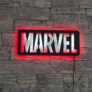 Könnte beinhalten: Ein schwarzes und weißes MARVEL-Logo-Schild mit roten LED-Lichtern um den Rand. Das Schild ist an einer grauen Steinwand montiert.