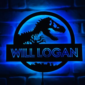 May include: A black, illuminated sign featuring a dinosaur skeleton silhouette within a circular frame. The sign includes the name "WILL LOGAN" in a blue, glowing font, set against a blue-lit brick wall. The design evokes a prehistoric theme.