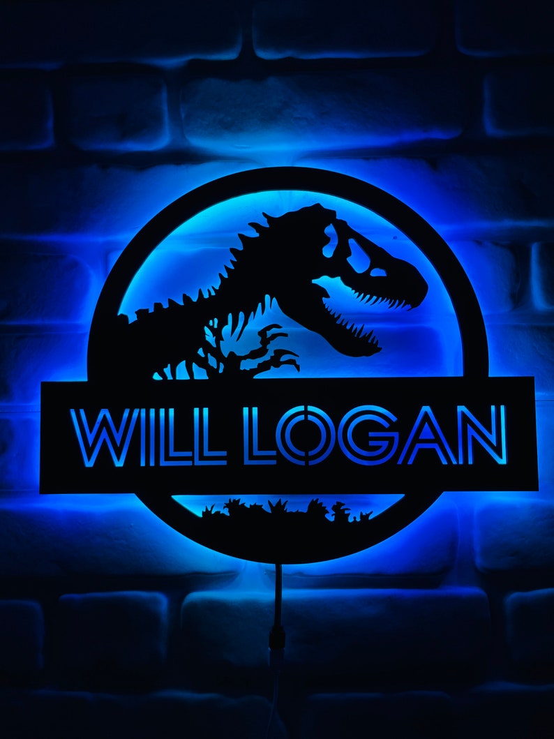 May include: A black, illuminated sign featuring a dinosaur skeleton silhouette within a circular frame. The sign includes the name "WILL LOGAN" in a blue, glowing font, set against a blue-lit brick wall. The design evokes a prehistoric theme.