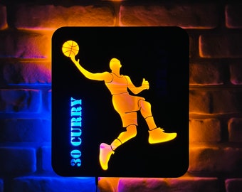 May include: Illuminated wall art featuring a basketball player silhouette in yellow against a black background. The player is mid-jump, holding a basketball. The text "30 CURRY" is vertically displayed. The art is framed by a square with blue and yellow lighting.