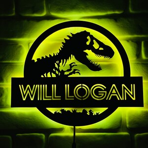 May include: Illuminated black sign featuring a dinosaur silhouette within a circular frame, with the text "WILL LOGAN" below. The sign is lit with a yellow glow against a brick wall background. A decorative item for a bedroom or game room.