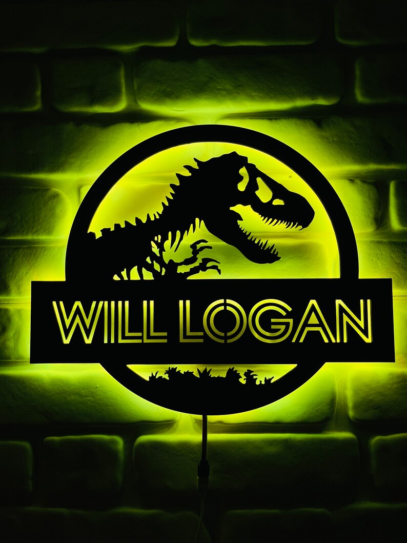 May include: Illuminated black sign featuring a dinosaur silhouette within a circular frame, with the text "WILL LOGAN" below. The sign is lit with a yellow glow against a brick wall background. A decorative item for a bedroom or game room.