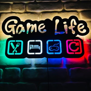 May include: A black neon sign with the words "Game Life" in white. The sign has four icons: a fork and knife, a bed, a video game controller, and a circle with an arrow. The icons are lit up in different colours: green, blue, and red.