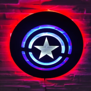 May include: A black circular wall light with a red and blue illuminated design resembling a superhero's shield. The shield features a white star in the center.