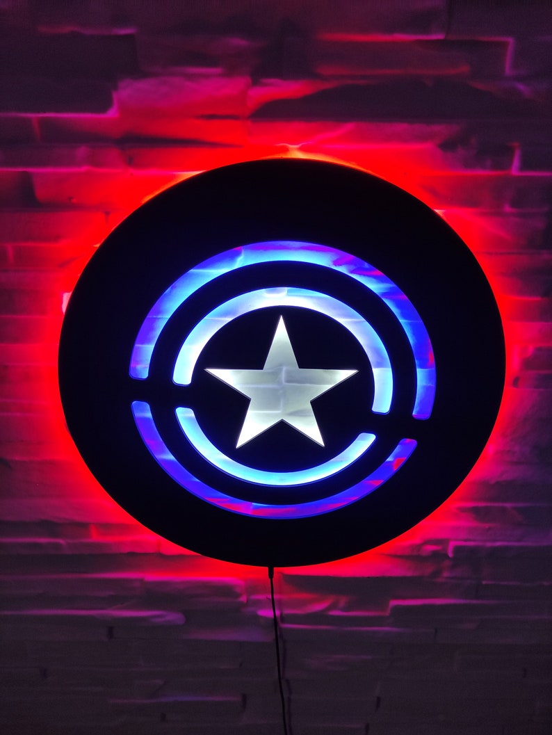 May include: A black circular wall light with a red and blue illuminated design resembling a superhero's shield. The shield features a white star in the center.