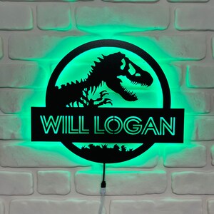 May include: Black dinosaur silhouette sign with the name "WILL LOGAN" illuminated by green LED lights. The sign is mounted on a white brick wall, creating a striking contrast. The design features a circular logo with a dinosaur skeleton.