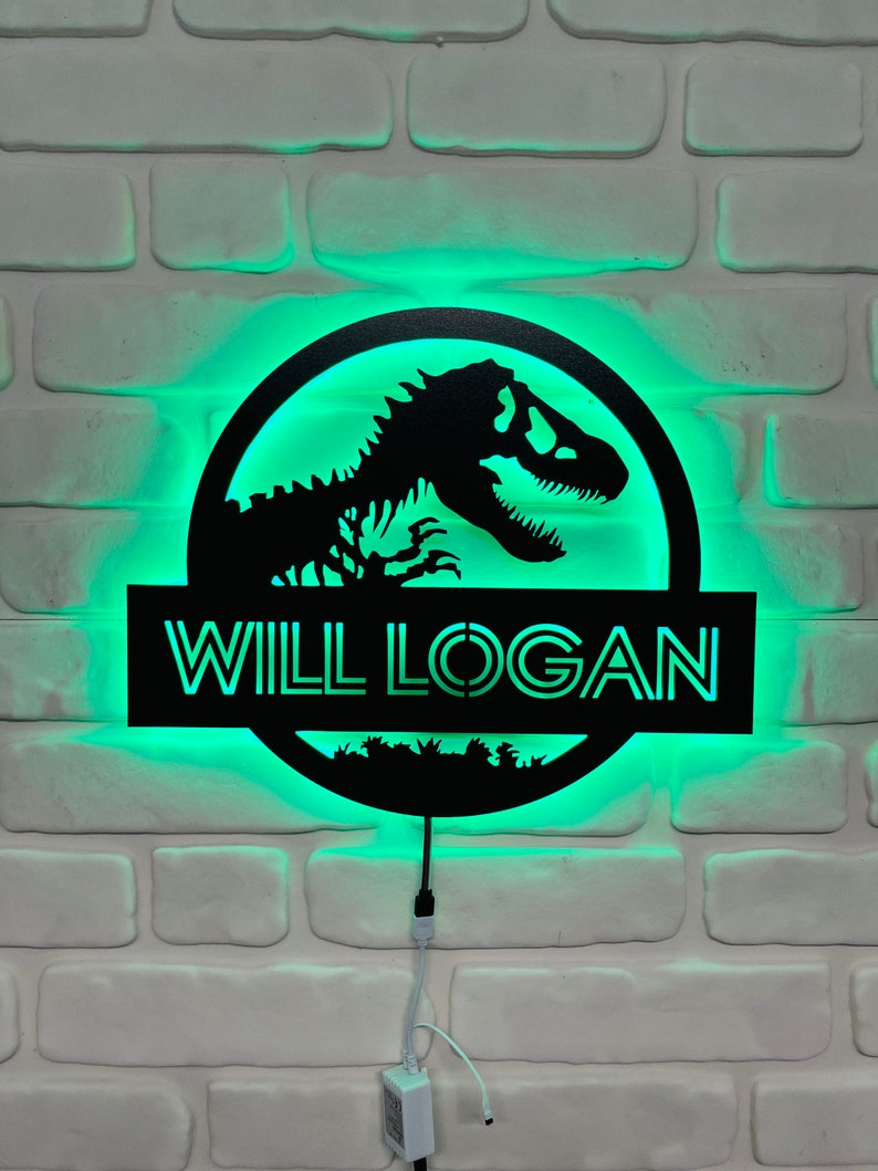 May include: Black dinosaur silhouette sign with the name "WILL LOGAN" illuminated by green LED lights. The sign is mounted on a white brick wall, creating a striking contrast. The design features a circular logo with a dinosaur skeleton.