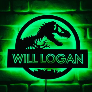 May include: A black, illuminated sign with the text "WILL LOGAN" and a dinosaur silhouette. The sign is circular with a rectangular base, and the background is a green-lit brick wall.