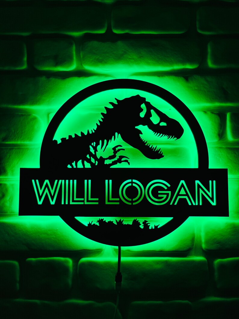 May include: A black, illuminated sign with the text "WILL LOGAN" and a dinosaur silhouette. The sign is circular with a rectangular base, and the background is a green-lit brick wall.