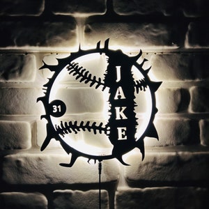 Pode incluir: Uma luminária de parede preta com tema de beisebol com o nome "JAKE" e o número "31" recortados. A luz tem a forma de uma bola de beisebol com costuras decorativas e uma borda pontiaguda. A luz é montada em uma parede de tijolos brancos.