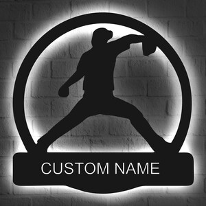 Pode incluir: Silhueta preta de metal de um jogador de beisebol lançando uma bola, com um círculo de luz atrás do jogador. A silhueta do jogador está montada em uma placa de metal preta com o texto "CUSTOM NAME" em branco.