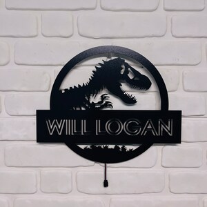 May include: Black metal sign featuring a dinosaur silhouette within a circular frame. The name "WILL LOGAN" is displayed below the dinosaur, with a small landscape design at the bottom. The sign is mounted on a white brick wall.