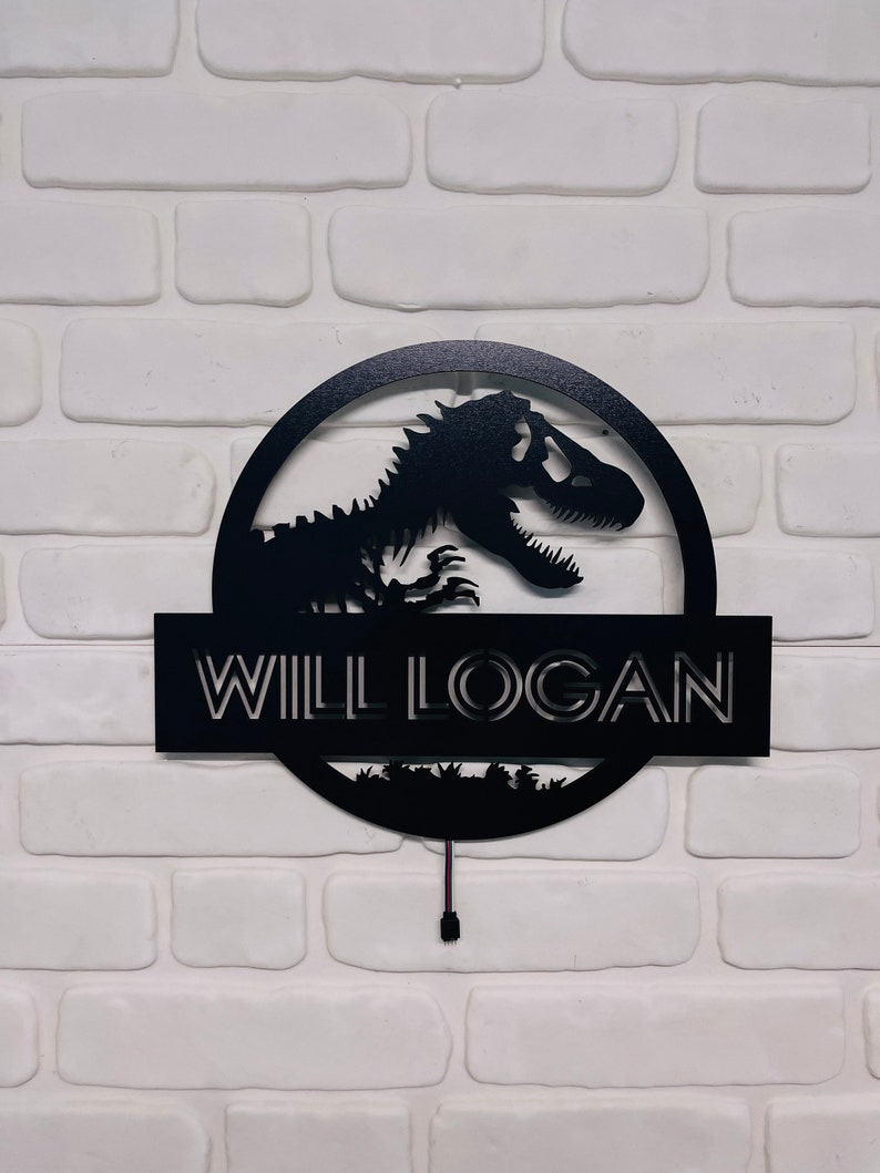 May include: Black metal sign featuring a dinosaur silhouette within a circular frame. The name "WILL LOGAN" is displayed below the dinosaur, with a small landscape design at the bottom. The sign is mounted on a white brick wall.