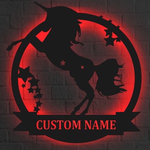 Pode incluir: Uma silhueta preta de metal de um unicórnio com um chifre e uma crina fluida, saltando dentro de um círculo com estrelas. O círculo tem uma faixa na parte inferior com o texto "CUSTOM NAME". O fundo é uma parede de tijolo.