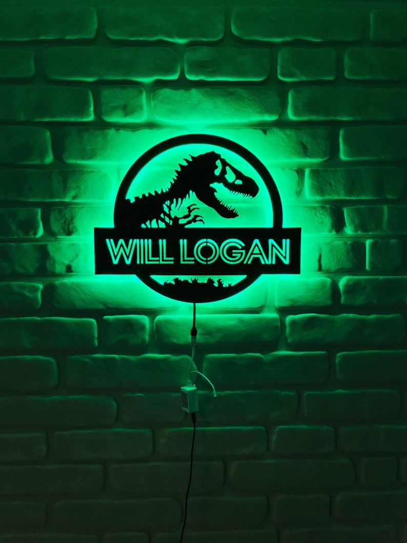 May include: A black, illuminated sign with the text "WILL LOGAN" and a dinosaur silhouette. The sign is lit with a green glow against a brick wall background. The sign is connected to a power source.