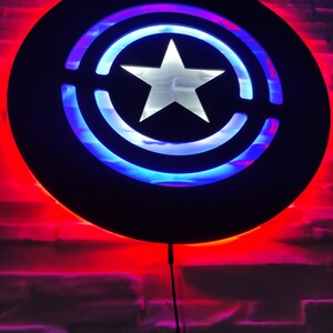 May include: A black circular wall light with a white star in the center. The light is surrounded by a blue and red ring. The light is on and illuminating the wall behind it.