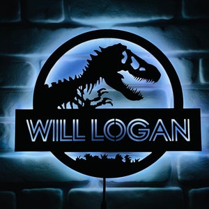 May include: A black, illuminated sign featuring a dinosaur skeleton silhouette within a circular frame. The name "WILL LOGAN" is displayed in white, block letters below the circle. The sign is mounted on a brick wall with a blue glow.