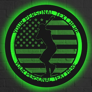 Pode incluir: Uma silhueta preta de metal de um jogador de beisebol balançando um taco, com um design de bandeira americana no fundo. A silhueta é cercada por um círculo com o texto "YOUR PERSONAL TEXT HERE" na parte superior e inferior.