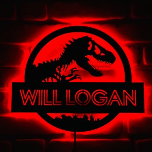 May include: A black illuminated sign with the name "WILL LOGAN" in red, featuring a dinosaur silhouette within a circular frame. The sign is mounted on a brick wall and emits a red glow.
