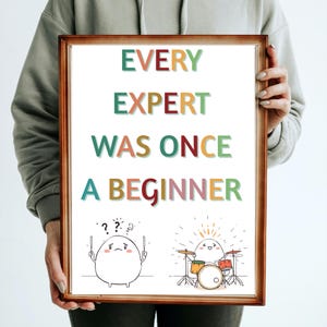 May include: A framed print with the text "EVERY EXPERT WAS ONCE A BEGINNER" in colorful letters. The print features two cartoon egg characters, one confused and the other playing drums. The frame is a warm brown color.