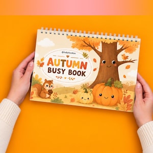 Op de afbeelding: Een spiraalgebonden Autumn Busy Book met een lichte kaft voorzien van een cartoonillustratie van een lachende boom, een eekhoorn met een eikel en twee pompoenen. De tekst luidt Autumn Busy Book met de merknaam babykookoo erboven.