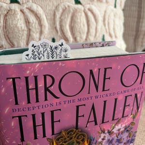 May include: A white floral bookmark with black line art of flowers and butterflies is placed in a book titled "Throne of the Fallen." The book has a pink cover with gold accents. A cream-colored pumpkin-shaped pillow is in the background.