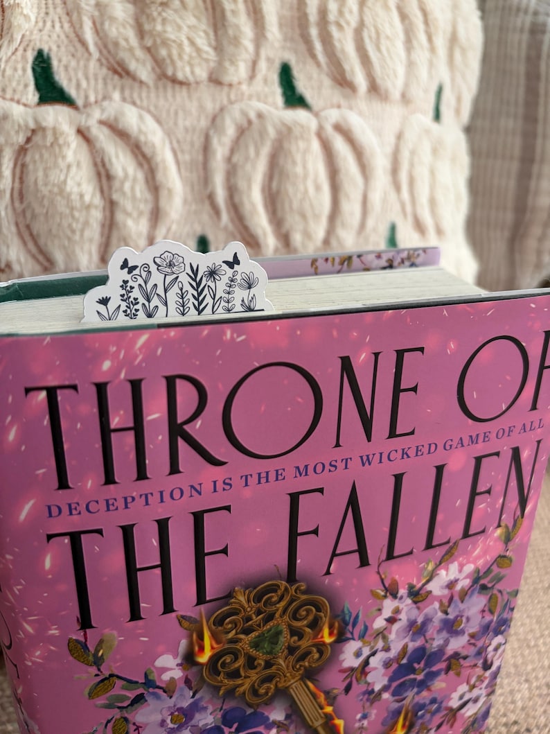 May include: A white floral bookmark with black line art of flowers and butterflies is placed in a book titled "Throne of the Fallen." The book has a pink cover with gold accents. A cream-colored pumpkin-shaped pillow is in the background.