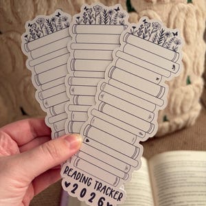May include: A stack of white reading tracker stickers with a floral design at the top and the text "Reading Tracker 2026". The stickers are held in a hand, with an open book in the background.