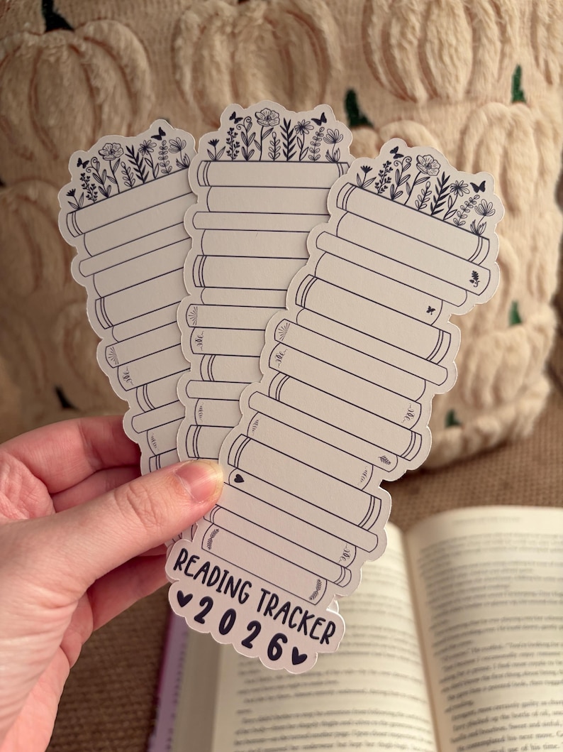 May include: A stack of white reading tracker stickers with a floral design at the top and the text "Reading Tracker 2026". The stickers are held in a hand, with an open book in the background.