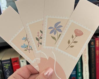 May include: Set of four floral bookmarks with a light pink background. Each bookmark features a different flower in a vintage-style stamp design. The flowers are a blue flower, a pink rose, a cluster of pink flowers, and a cluster of blue flowers.
