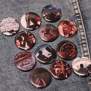 May include: A collection of 12 round pins with various images and text related to the Hunger Games. The pins are red, black, and white with images of characters, symbols, and locations from the book series. The text on the pins includes "If we burn, you burn with us", "The Hunger Games", "District 12", "Mellark Bakery", "Victor Village", and "Capitol"