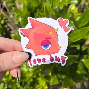 May include: A red and white sticker featuring a cartoon dinosaur with a blue eye and the text "love bug". Two pink hearts are in the top right corner of the sticker.