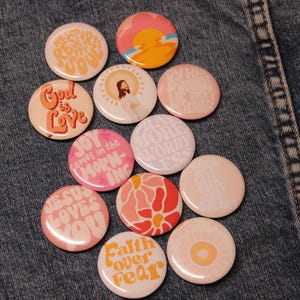 May include: A collection of round, colorful pinback buttons with various designs and text. The buttons feature phrases like "God is Love," "Jesus Loves You," and "Faith Over Fear." The color palette includes pink, orange, and white.