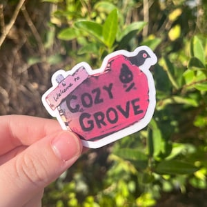 May include: A pink and white sticker with a bird on top. The sticker says "Welcome to Cozy Grove" with a stylized campfire graphic.