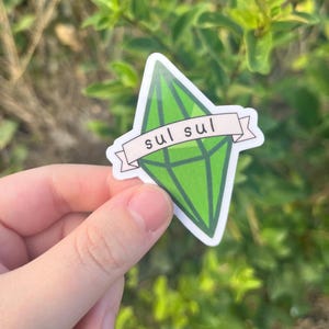 以下が含まれることがあります： 白いバナーに「sul sul」と書かれた緑色のダイヤモンド型のステッカー。このステッカーは、ビデオゲーム「ザ・シムズ」の象徴的なシンボルです。