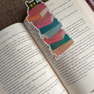 Peut inclure: Un marque-page coloré représentant une pile de livres avec un chat noir qui regarde par-dessus. Le marque-page est placé dans un livre ouvert avec du texte visible sur les pages.