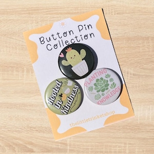 May include: A set of three round button pins on a white and yellow card. The pins feature a cactus, a succulent, and the phrases "Rooted in Kindness" and "Planting Knowledge". The card reads "Button Pin Collection".