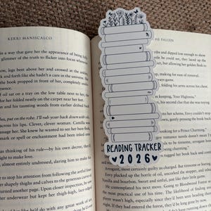 May include: A white bookmark with a stack of books design, topped with floral illustrations. The bookmark has the text "READING TRACKER 2026" in black. The bookmark is placed in an open book.