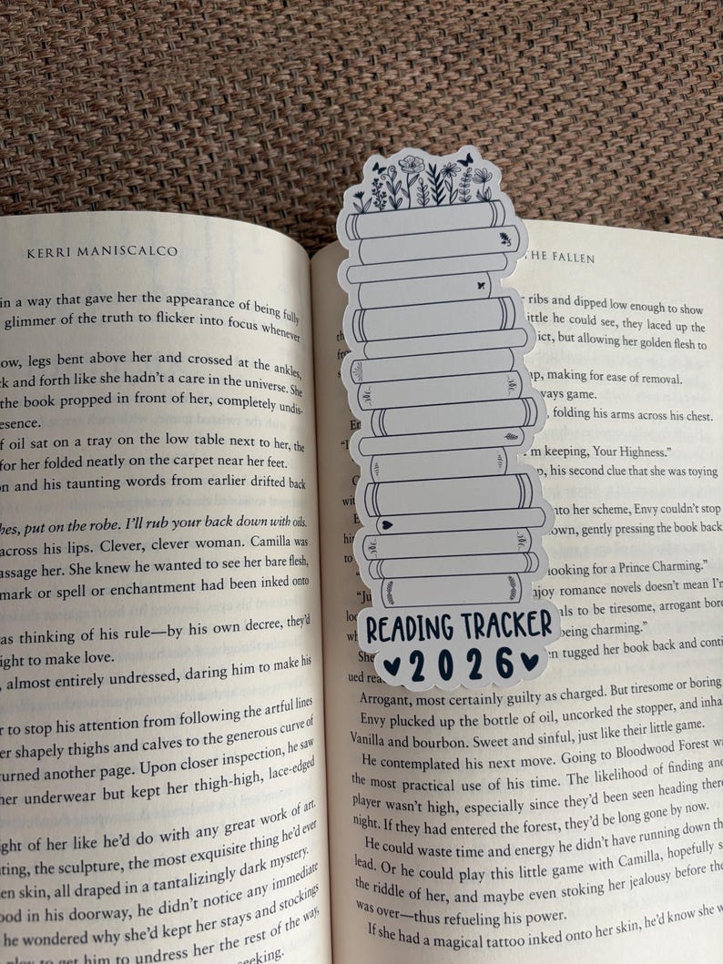 May include: A white bookmark with a stack of books design, topped with floral illustrations. The bookmark has the text "READING TRACKER 2026" in black. The bookmark is placed in an open book.