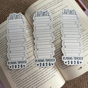 May include: Three white and black reading tracker bookmarks with a stack of books design, topped with flowers and butterflies. Each bookmark has the text "READING TRACKER 2026" with a heart symbol. The bookmarks are placed on an open book.
