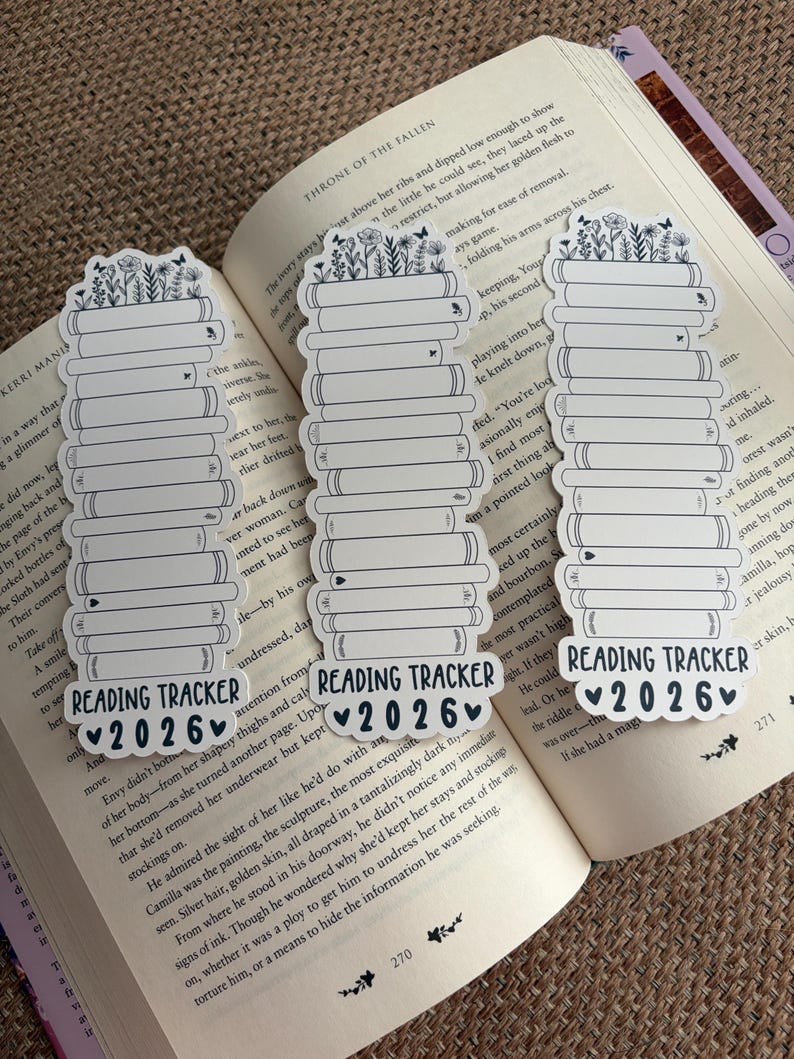 May include: Three white and black reading tracker bookmarks with a stack of books design, topped with flowers and butterflies. Each bookmark has the text "READING TRACKER 2026" with a heart symbol. The bookmarks are placed on an open book.