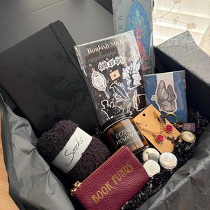 Puede incluir: Una caja de regalo negra que contiene un cuaderno negro con un diseño de cuervo, una hoja de pegatinas góticas, un marcapáginas, una pegatina de mariposa, una vela etiquetada October Mourning, calcetines negros suaves, un monedero burdeos con el texto Book Funds y joyas.