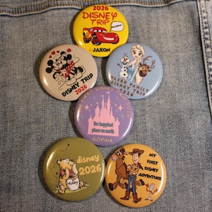 May include: A collection of seven round Disney-themed buttons. The buttons feature characters like Mickey and Minnie Mouse, Lightning McQueen, Winnie the Pooh, Elsa, and Woody. Each button has the text "Disney Trip 2026" or "My First Disney Adventure".