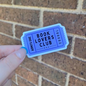 May include: A blue and white sticker in the shape of a ticket. The sticker reads "BOOK LOVERS CLUB" in bold, white text. The words "ADMIT ONE" are on the left side, and the number "100002" is on the right.