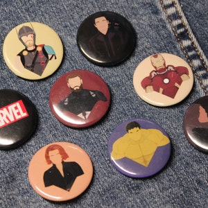 May include: Eight round buttons with different Marvel characters on them. The buttons are on a blue denim background. The characters include Iron Man, Captain America, Thor, Hulk, Black Widow, Spider-Man, and two others. One button says "MARVEL" in red letters on a black background.