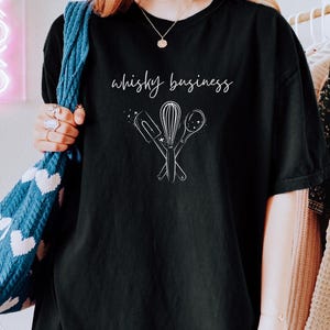 Pode incluir: Uma t-shirt preta com um gráfico de linhas brancas de uma espátula, um batedor e uma colher de pau cruzados. Acima dos utensílios de cozinha, o texto "whisky business" está impresso numa fonte cursiva branca de estilo manuscrito.